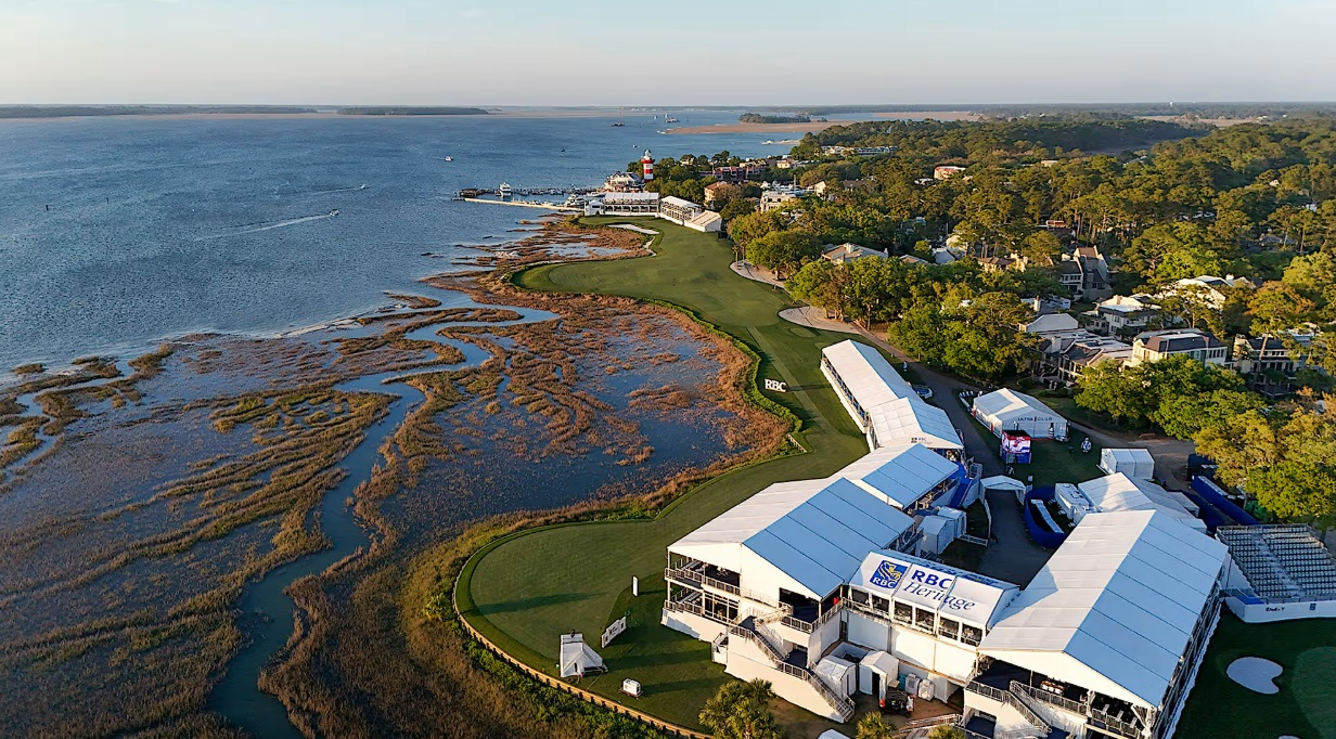 High-angle aerial view of Harbour Town Golf Links featuring lush green fairways alongside tidal marshlands and open blue water, with white event marquees branded RBC Heritage and the iconic red-and-white striped lighthouse visible in the distance.